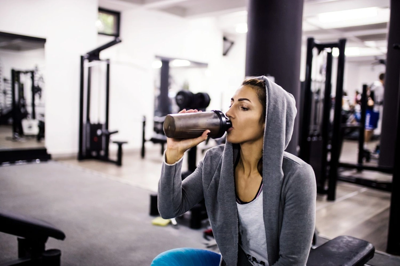 Person in a gym wearing a hoodie drinks a protein shake from a shaker bottle while sitting on a bench